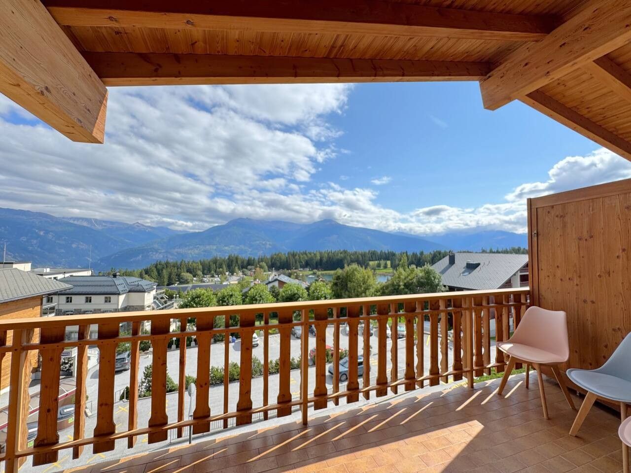 Un balcón con barandilla de madera, dos sillas y vistas a un paisaje montañoso con edificios al fondo.