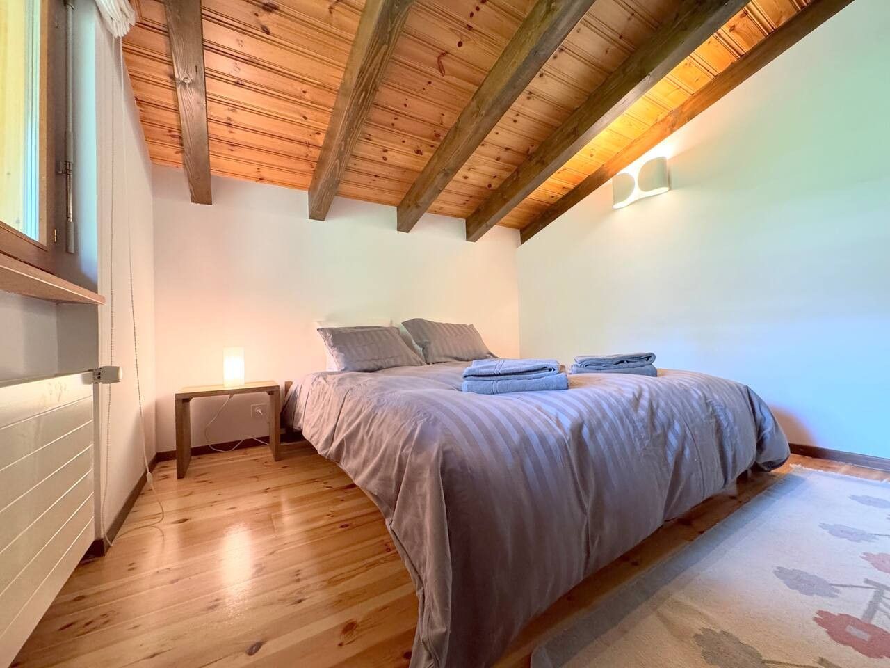 Ein Schlafzimmer mit Holzboden, einem Bett mit grauer Bettwäsche, einem Nachttisch mit Lampe und einem Fenster.