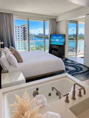 Ein Hotelzimmer mit einem großen Bett, einem Fernseher, einem Balkon mit Blick auf die Stadt und das Wasser sowie einer Badewanne mit Handtüchern.