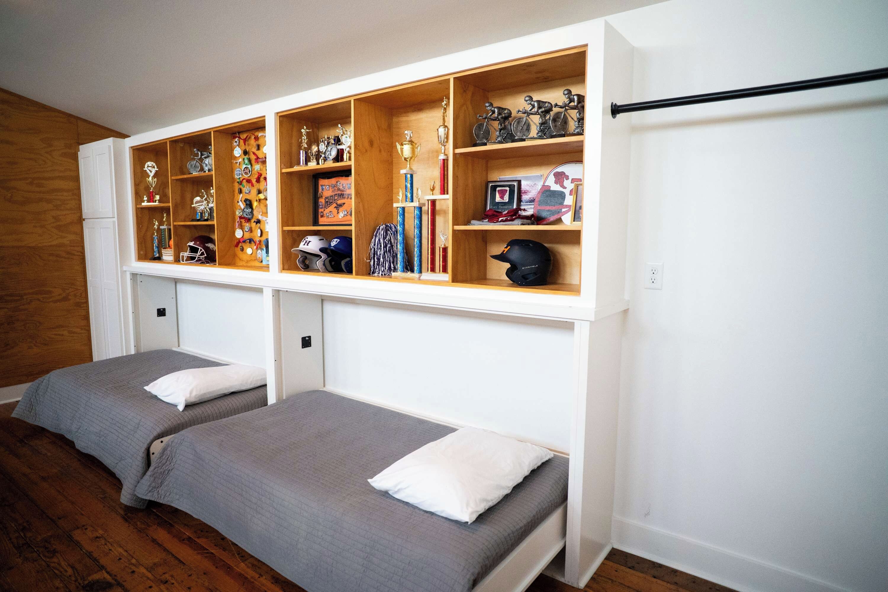 A bedroom with two single beds, a wooden shelf unit above with various trophies and helmets, and a white wall.