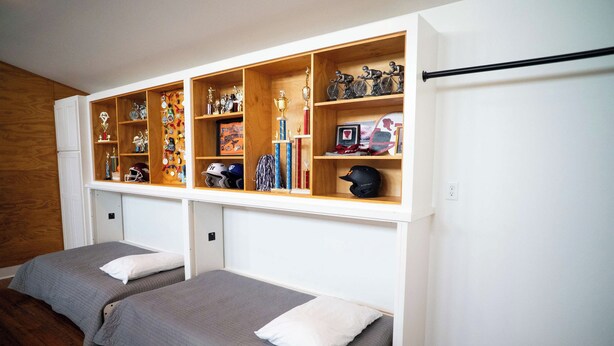A bedroom with two single beds, a wooden shelf unit above with various trophies and helmets, and a white wall.
