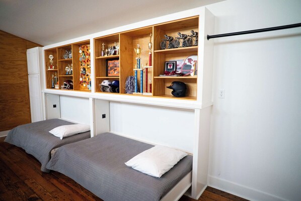 A bedroom with two single beds, a wooden shelf unit above with various trophies and helmets, and a white wall.