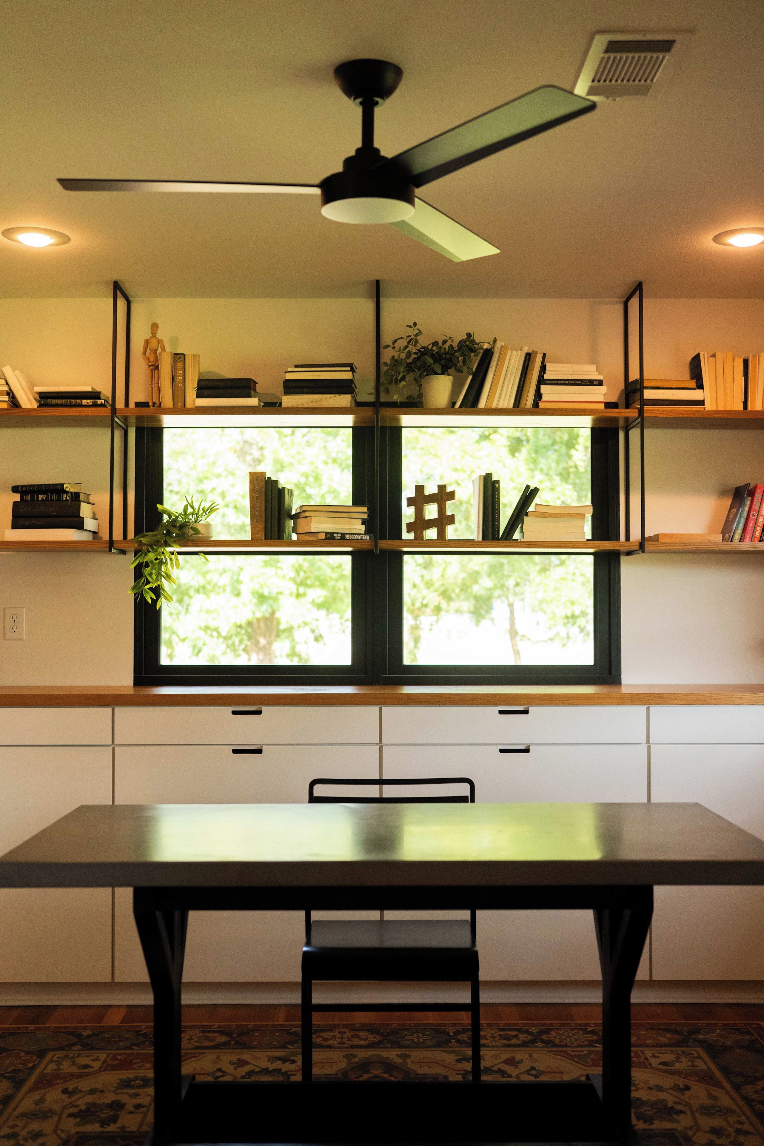 Una habitación con una mesa de comedor, sillas, un ventilador de techo y estantes con libros y objetos decorativos.