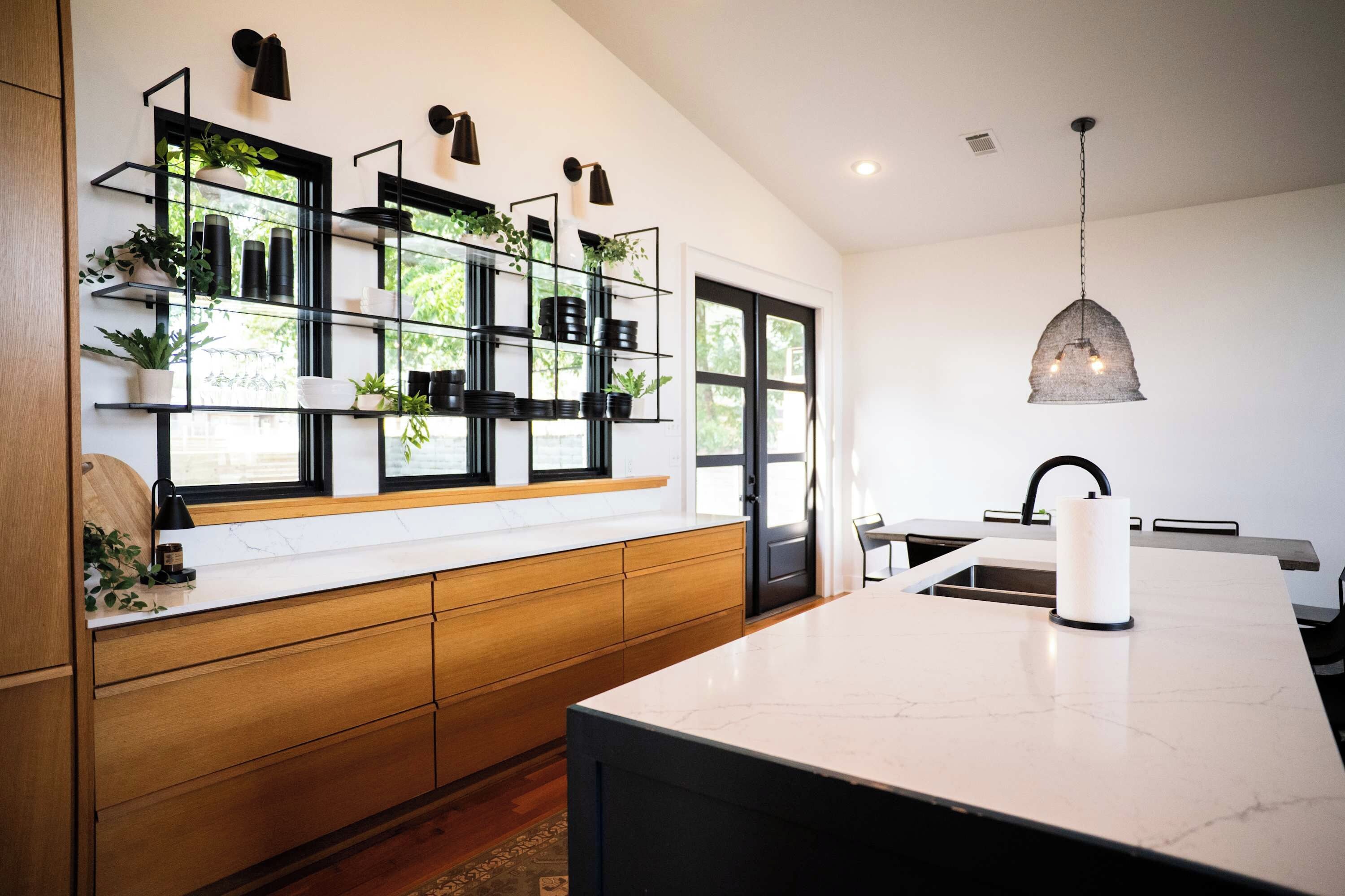 A modern kitchen with a marble countertop, wooden cabinets, and a built-in shelf unit with potted plants.