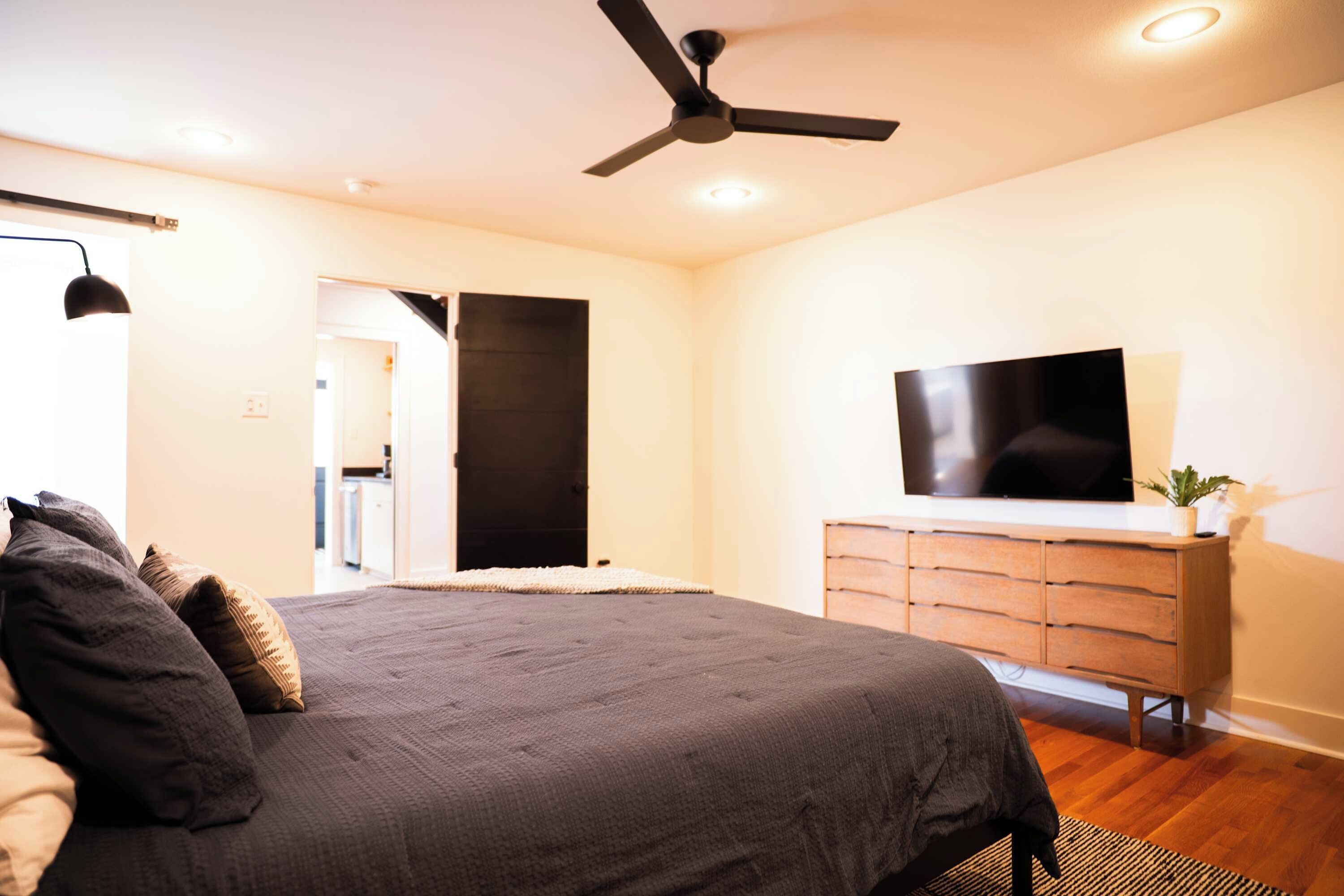 A bedroom with a bed, a ceiling fan, a TV on a dresser, and a door leading to another room.