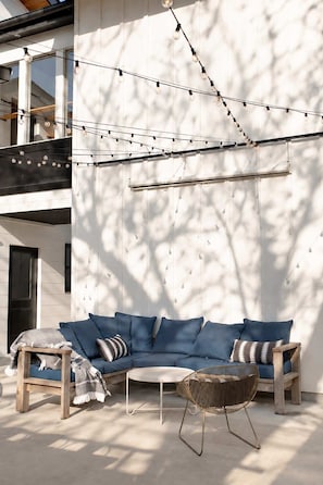 A modern outdoor seating area with a sofa and chairs, string lights, and a white wall. - Mid Mod Lodge - Patio - Office - 0.5 mi UCA (Conway)