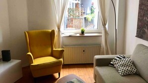 Ein Zimmer mit einem gelben Sessel, einem grauen Sofa, einem Tisch mit einem Buch, einem Heizkörper und einem Fenster mit Vorhängen.