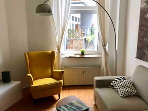 Une pièce avec un fauteuil jaune, un canapé gris, une table avec un livre, un radiateur et une fenêtre avec des rideaux.