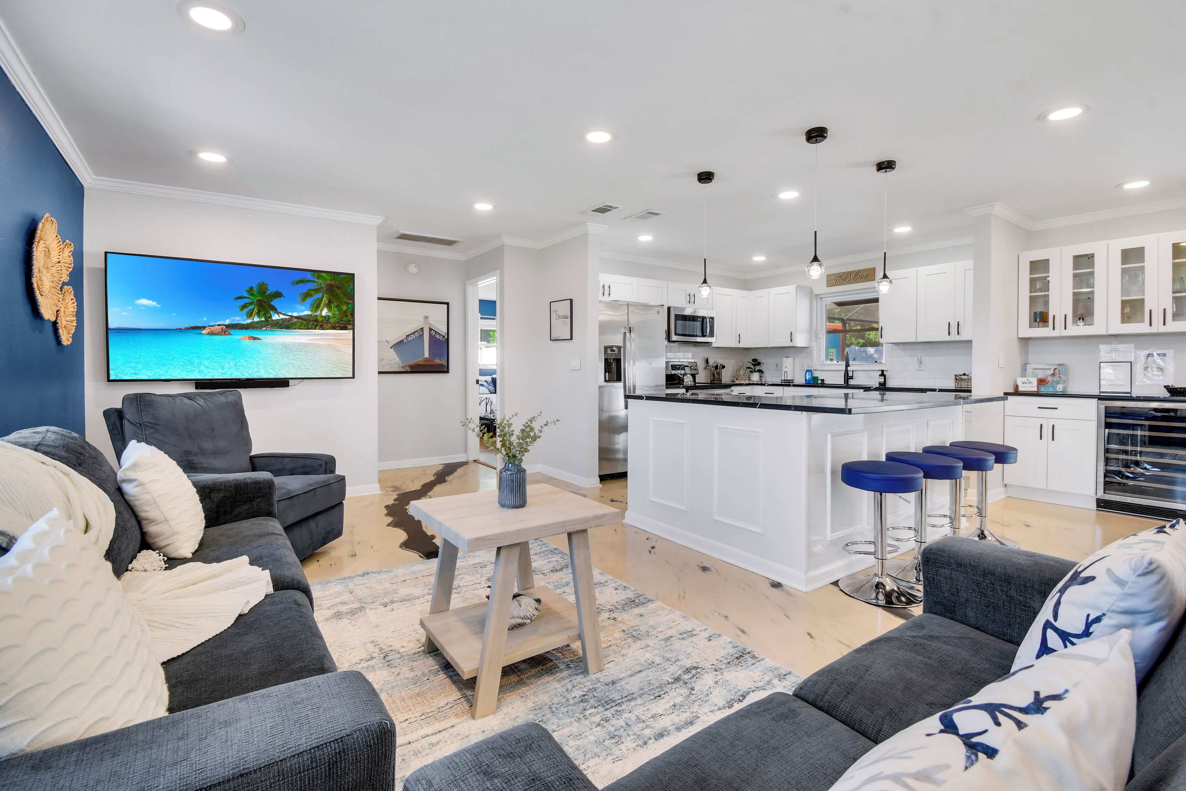 A modern living room with a flat-screen TV, grey sofas, a coffee table, and a kitchen with white cabinets and a bar area.