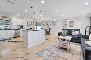 A modern kitchen with white cabinetry, a central island, and a dining area with a grey sofa and a coffee table. - Heated Pool w/ Hot Tub • 10 min to Beaches • Pinellas Trail Access (Largo)