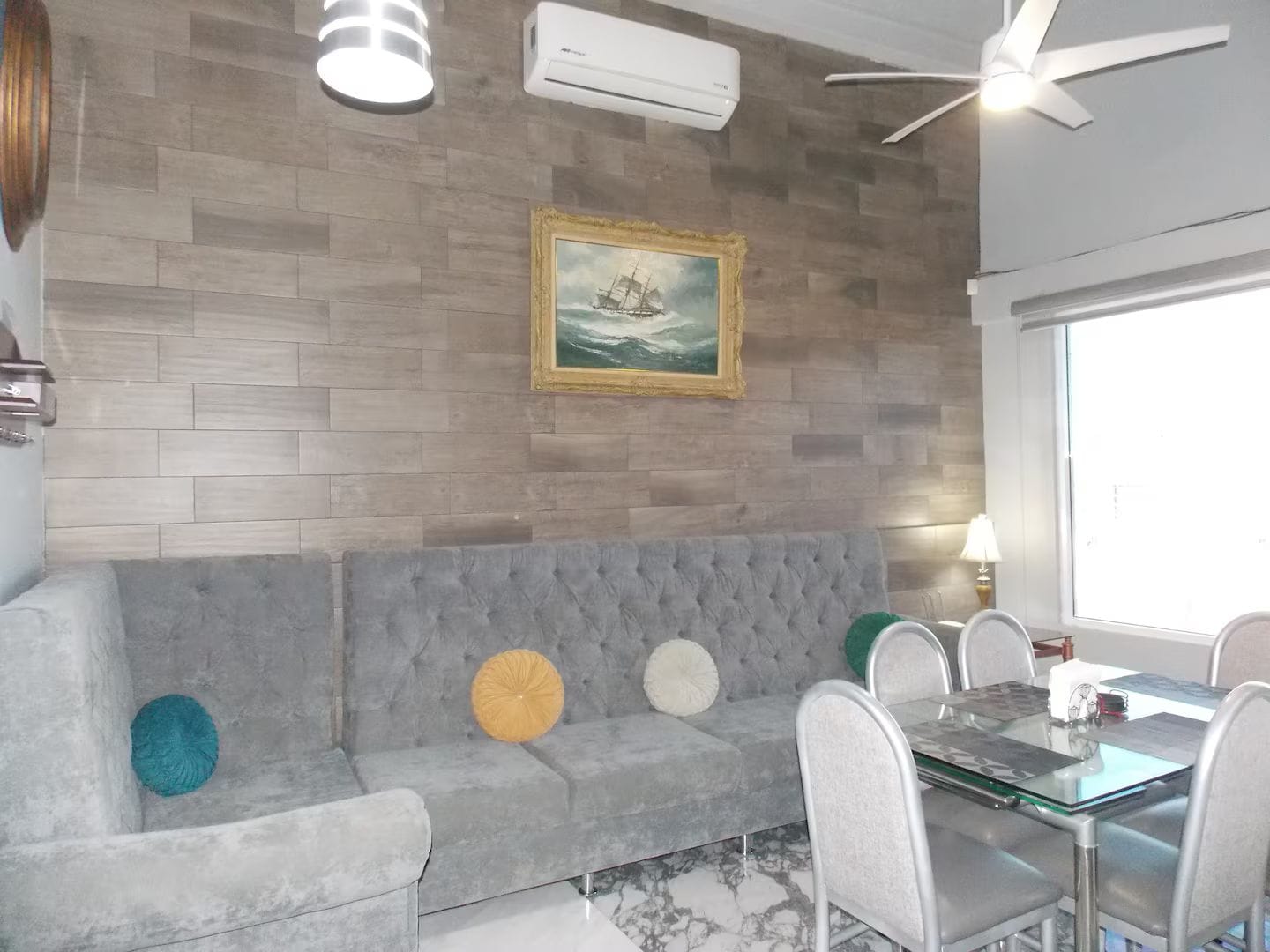 A modern living room with a grey sofa, a dining table with chairs, a ceiling fan, and an air conditioning unit.