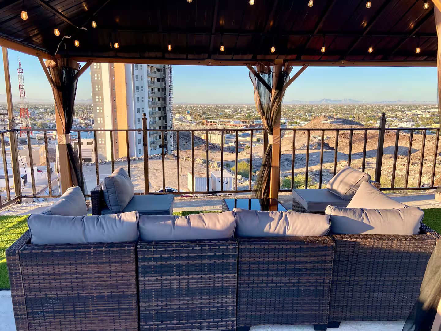 A covered outdoor seating area with wicker furniture and cushions, overlooking a cityscape with buildings and construction.