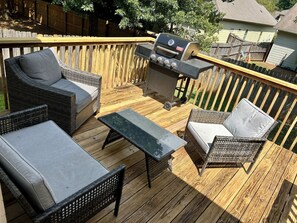 Una terraza de madera con mobiliario de mimbre, una parrilla y vistas a una zona residencial.