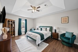 A bedroom with a bed, a chair, a television, a ceiling fan, and a window with curtains.