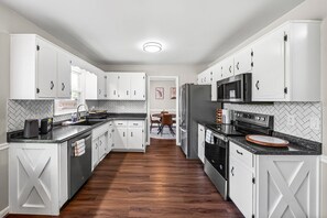 A modern kitchen with white cabinetry, a central island, and a dining area in the background. - Relax & Recharge  Cozy 2BR Near Fort Campbell (Clarksville)