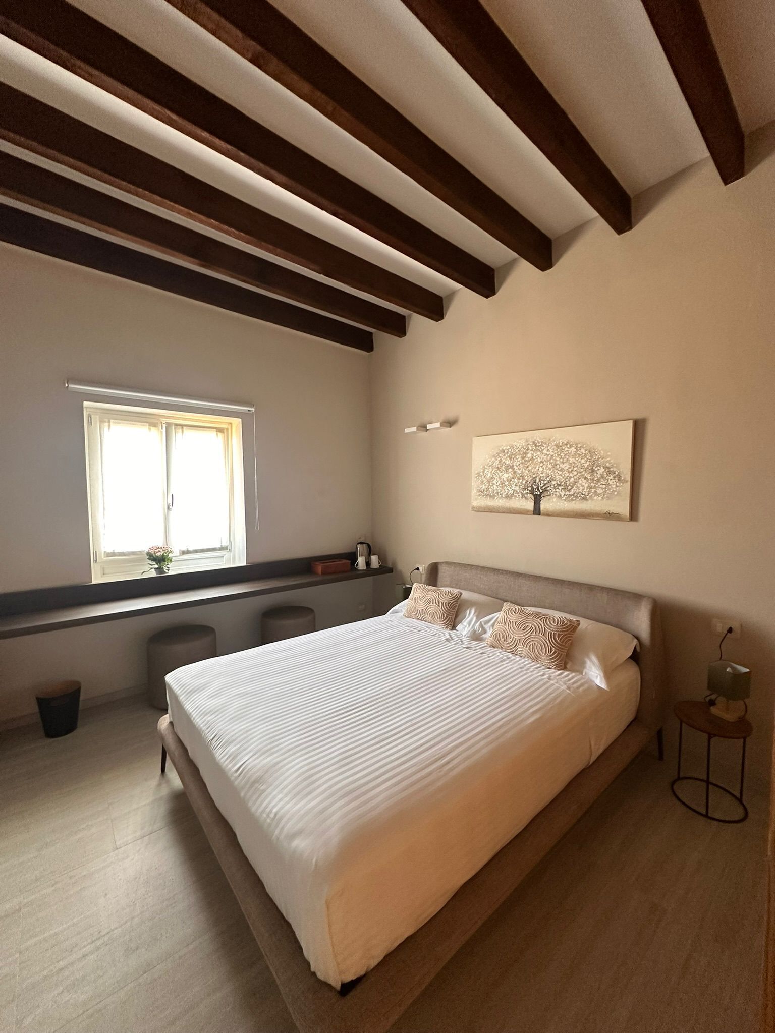 Un dormitorio con una cama grande, una ventana, un cuadro y una mesita.