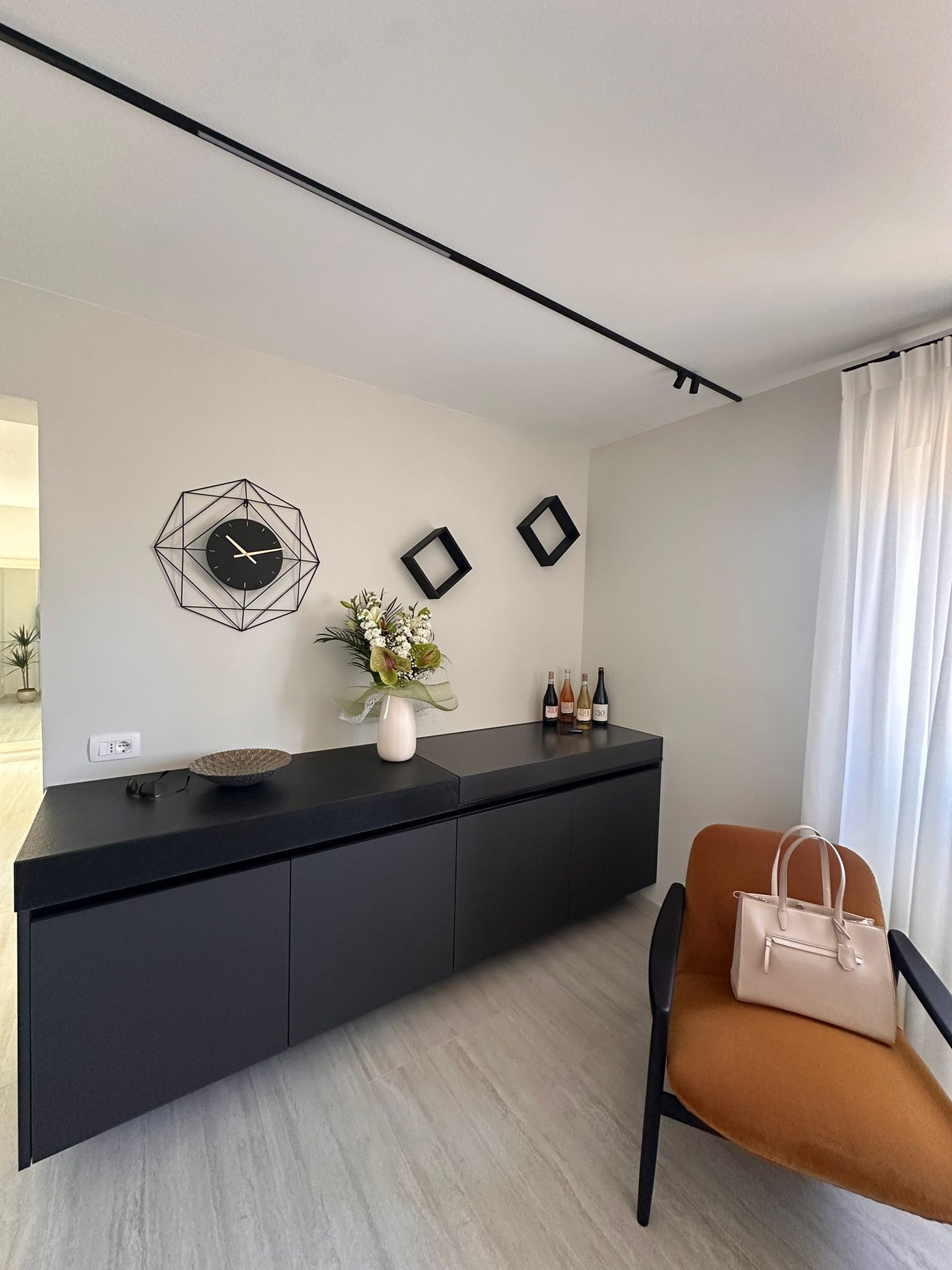 Una sala moderna con una mesa consola negra, un reloj geométrico, cuadros enmarcados, un florero con flores y una silla marrón con un bolso de mano.