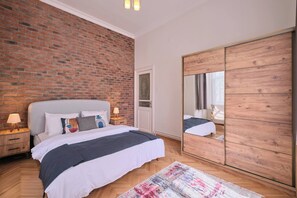 Ein Schlafzimmer mit Backsteinwand, einem Bett mit grauem Kopfteil, einem Holzschrank und einem Spiegel.