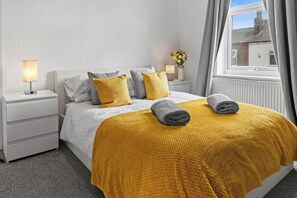 A neatly made bed with a white headboard and a mustard-colored blanket, two pillows, a bedside lamp, and a window with curtains. - Spacious 3BR Home | 15 Mins to Manchester (Swinton)