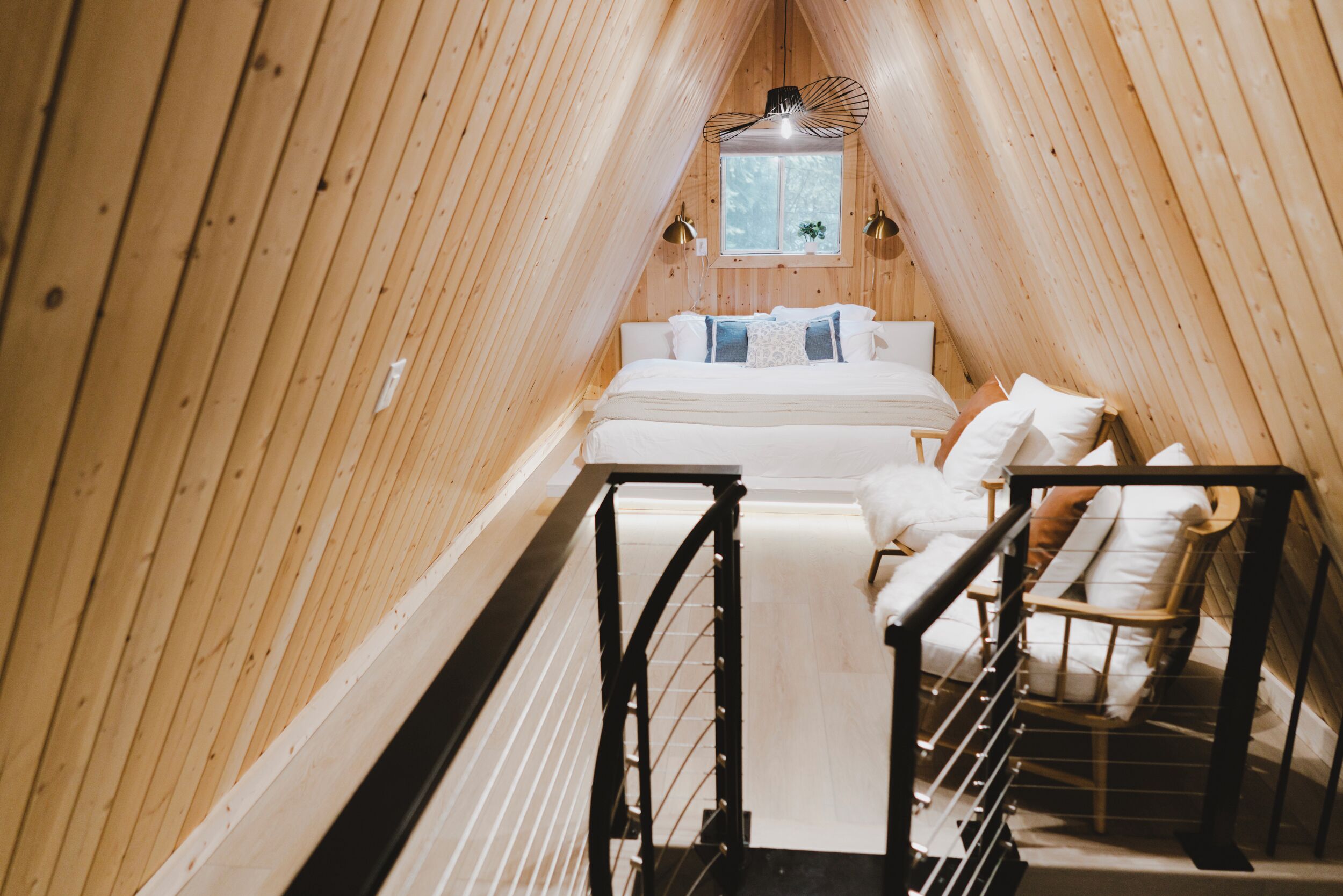 A narrow, wooden-paneled attic bedroom with a bed, two chairs, and a small window.