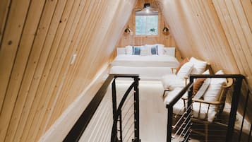 A narrow, wooden-paneled attic bedroom with a bed, two chairs, and a small window.