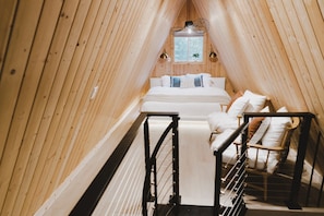 Un dormitorio abuhardillado estrecho con paneles de madera, una cama, dos sillas y un ventanal pequeño.
