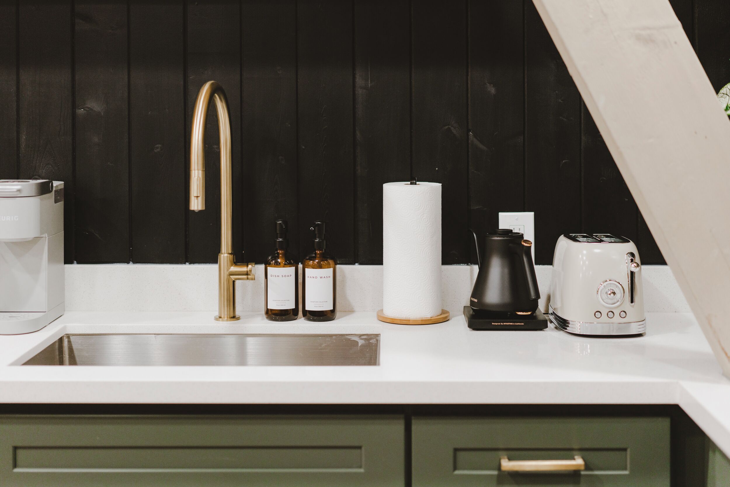 A kitchen with a stainless steel sink, a gold faucet, a coffee maker, a kettle, and two bottles of dish soap.