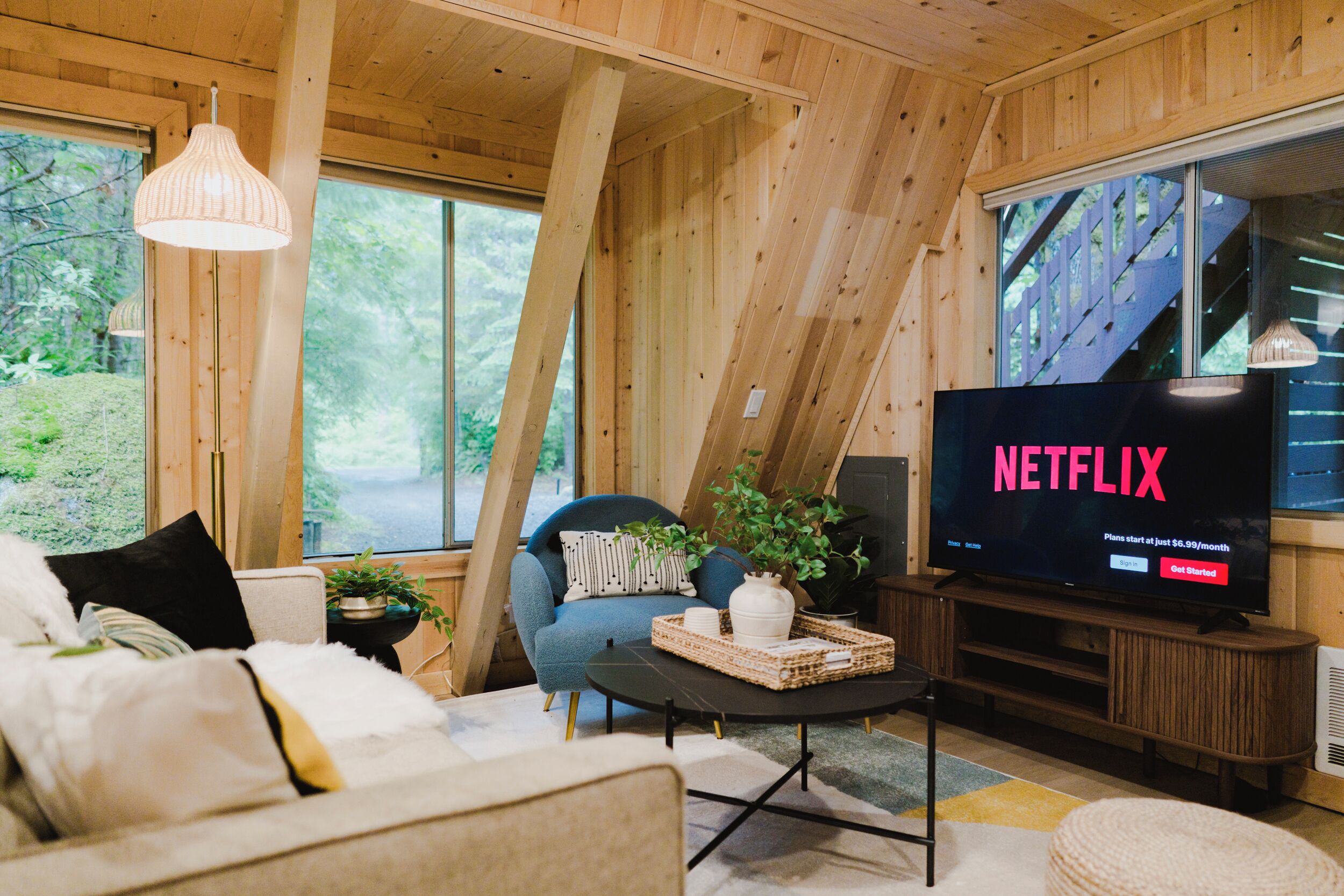 Un salon chaleureux avec un grand téléviseur affichant le logo de Netflix, des murs en bois, un canapé bleu et une fenêtre donnant sur un paysage verdoyant.