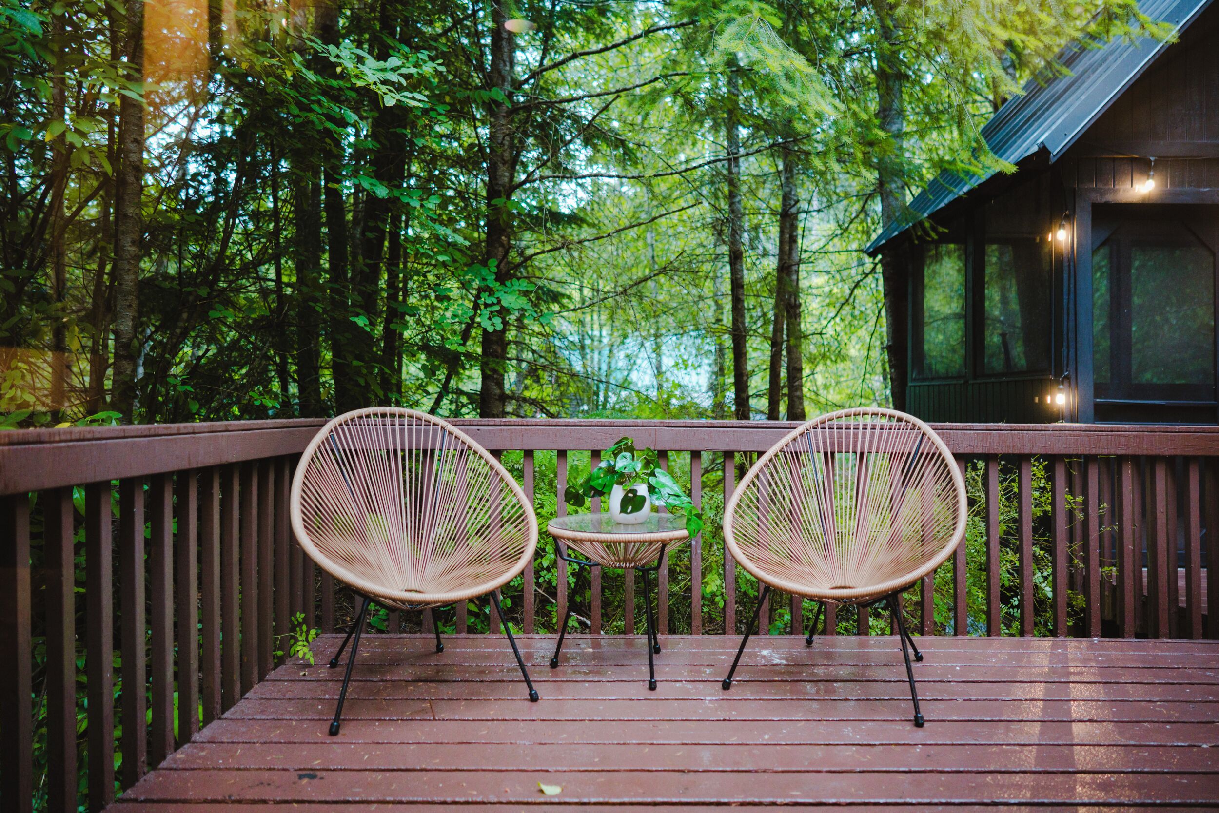 A wooden deck with two wicker chairs and a small table, surrounded by dense forest.