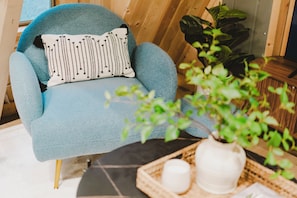 Un sillón azul con un cojín estampado, una planta en maceta y una bandeja con un jarrón blanco y una vela.