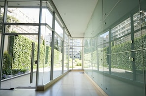 Un pasillo moderno con paredes de vidrio, piso de baldosas y vistas al exterior con zonas verdes y edificios.