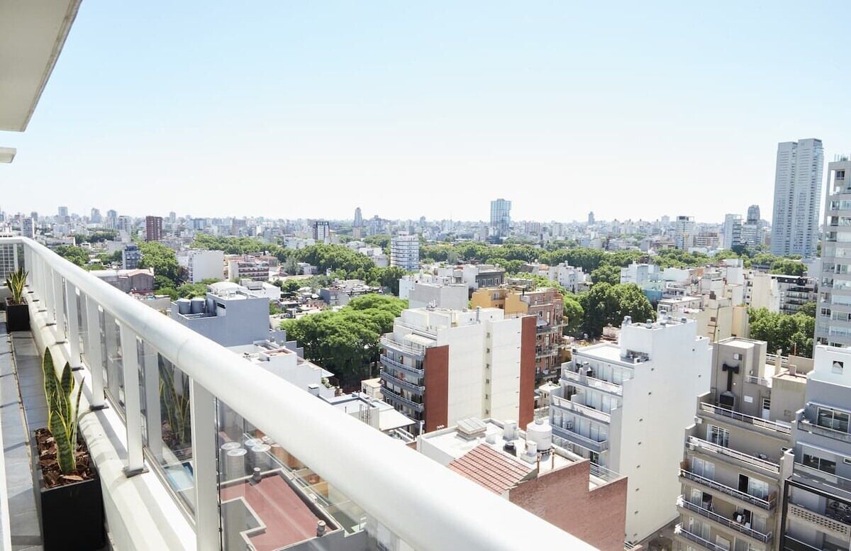 A cityscape with high-rise buildings, a clear sky, and a balcony with potted plants.