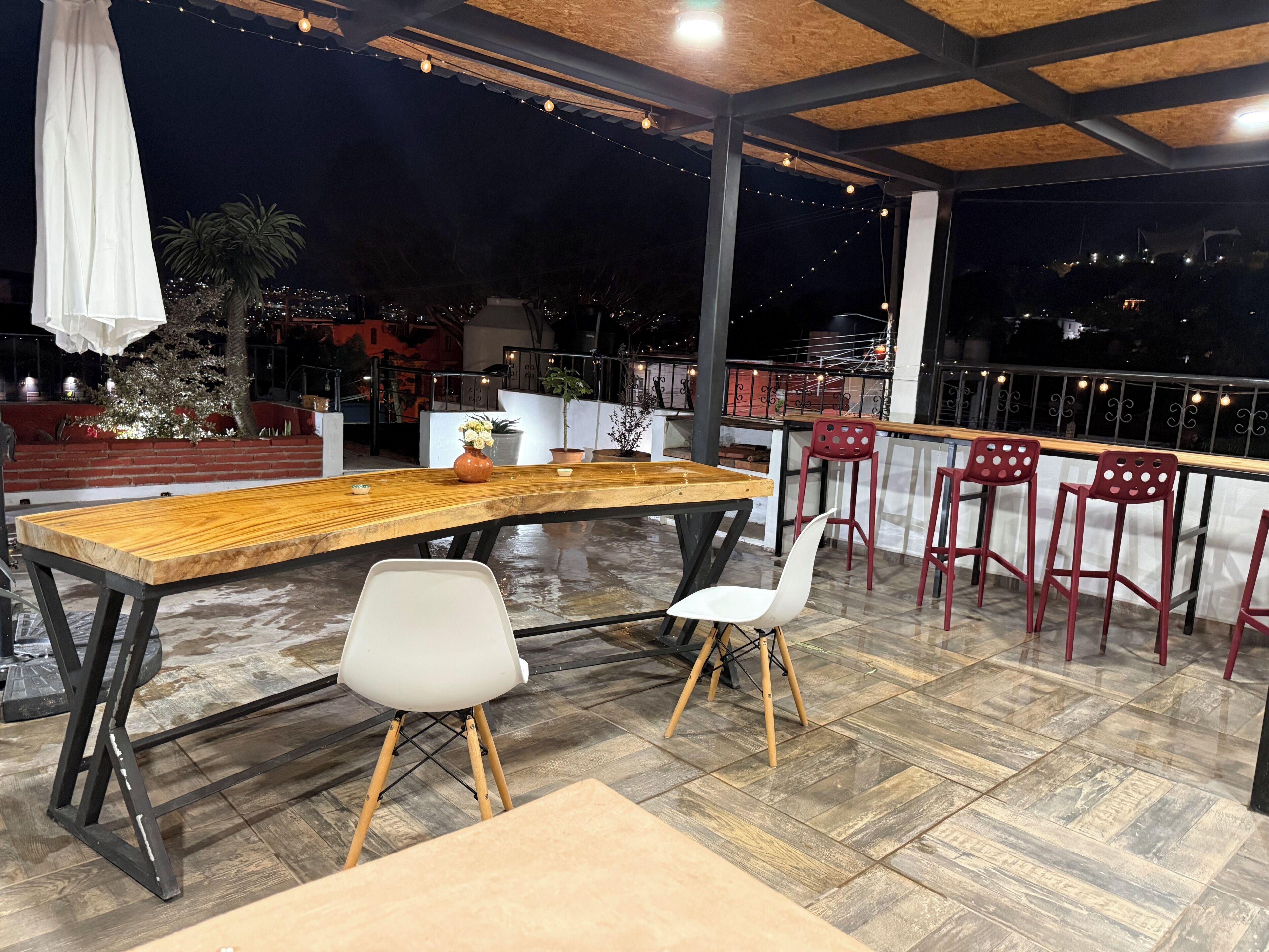 A rooftop terrace with a wooden table, two chairs, and a railing overlooking a cityscape at night.