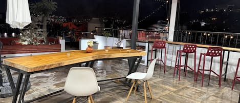 Una terraza en la azotea con una mesa de madera, dos sillas y una barandilla con vistas a la ciudad de noche.