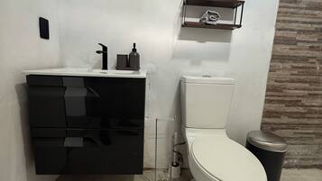 Un baño moderno con inodoro blanco, tocador negro y un pequeño estante sobre el inodoro.
