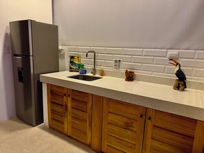 Uma cozinha com geladeira de aço inoxidável, pia com torneira, armários de madeira e bancada com diversos objetos, incluindo uma estatueta de pássaro e uma placa.