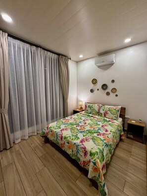 Une chambre à coucher avec une couvre-lit à motifs floraux, un plancher en bois, un appareil de climatisation et une table de chevet avec une lampe.