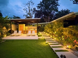 Un espace extérieur moderne aménagé avec un coin salon, une pergola, un bar et un chemin bordé de lumières.