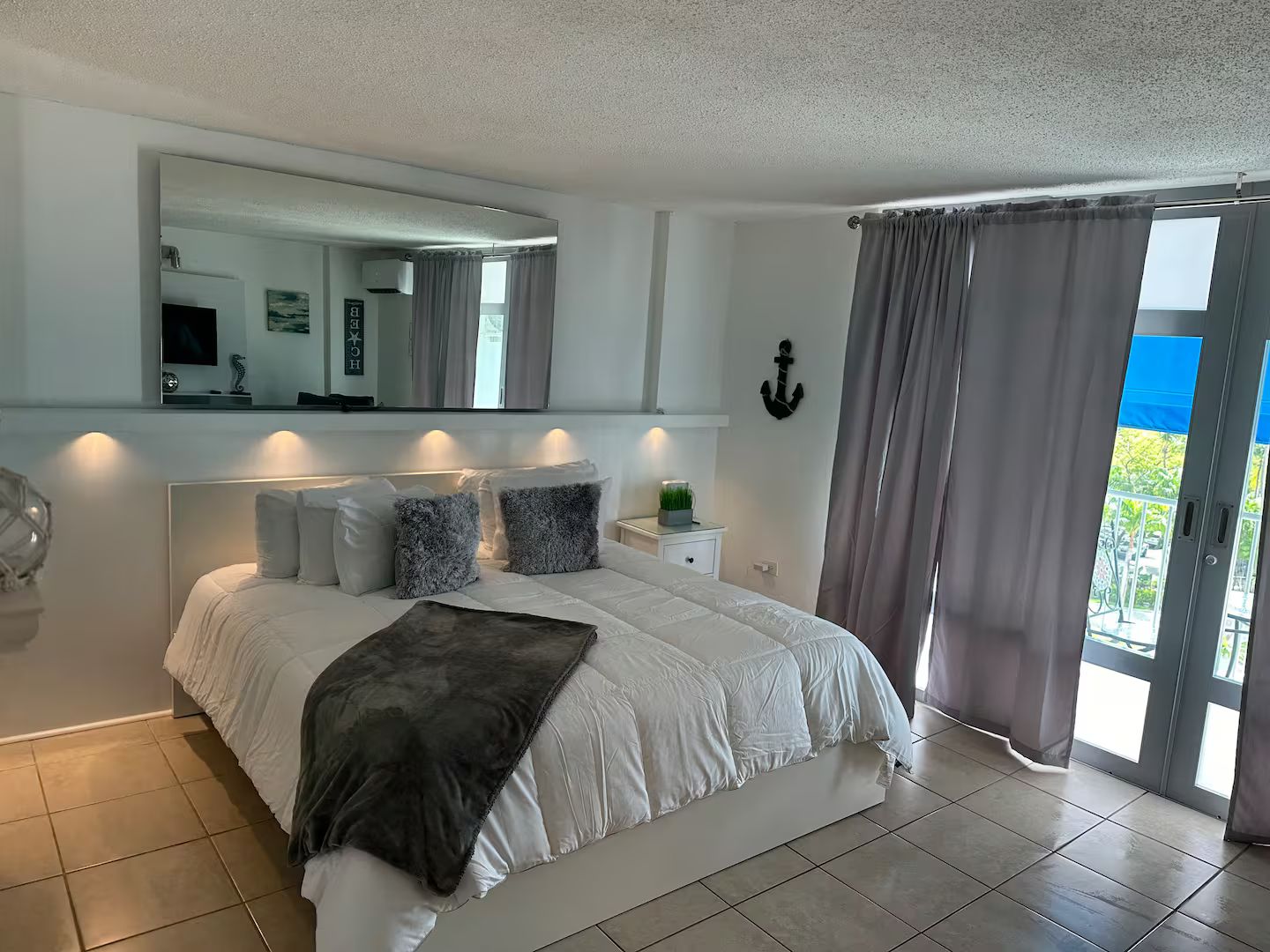 A bedroom with a large bed, a bedside table with a plant, a mirror, and a view of the outdoors.
