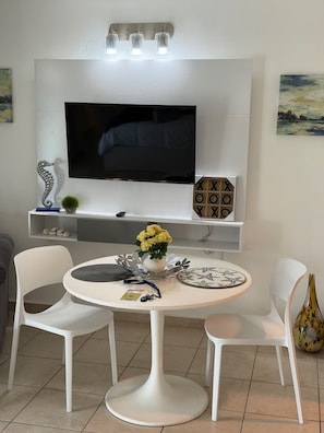 Ein modernes Wohnzimmer mit einem Flachbildfernseher, weißen Stühlen, einem runden Tisch und Dekorationselementen.