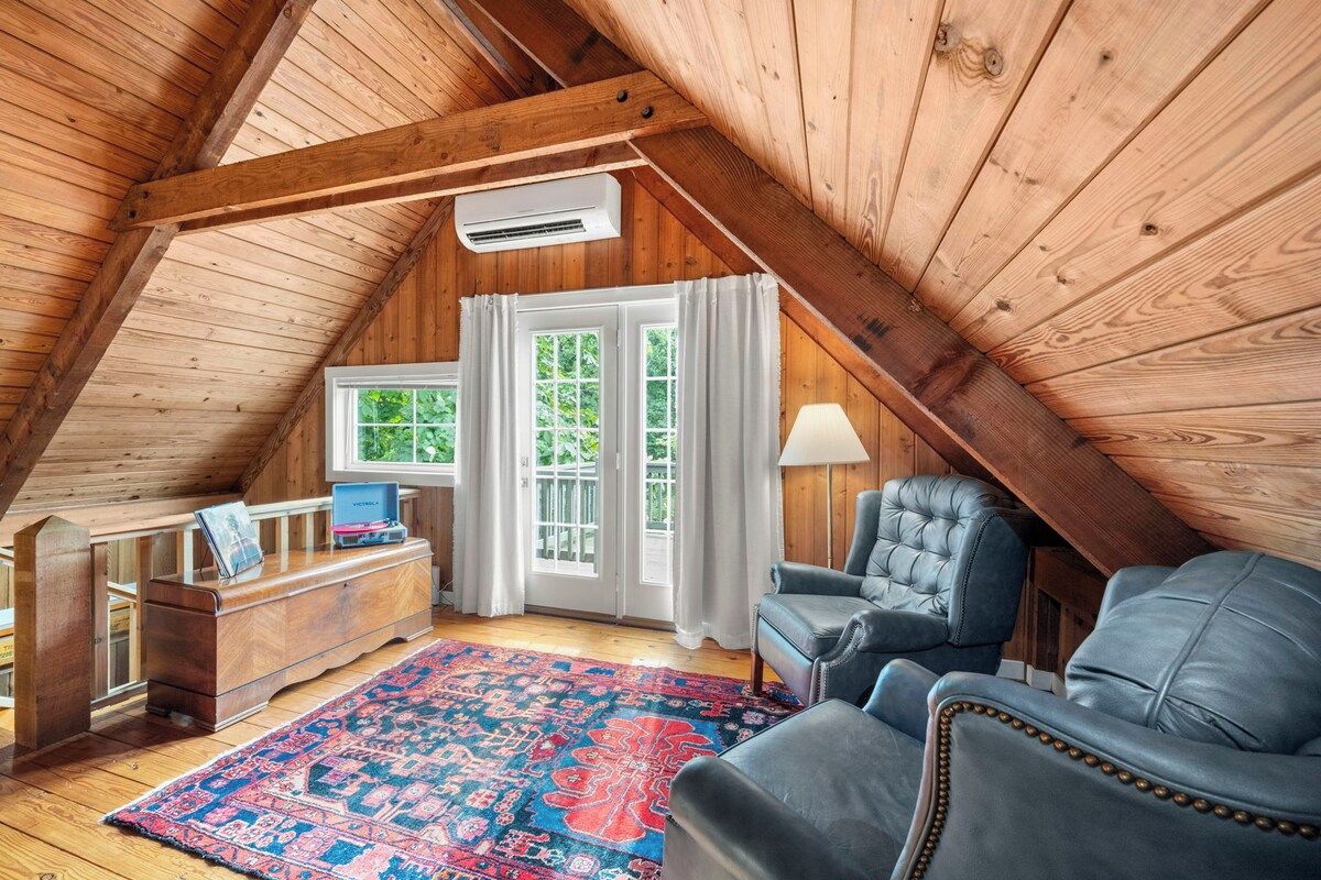A cozy attic room with wooden beams, a desk, two chairs, a rug, and a window with curtains.