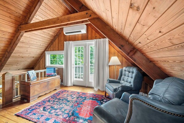 A cozy attic room with wooden beams, a desk, two chairs, a rug, and a window with curtains.