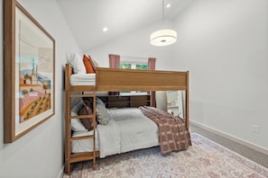 A bunk bed with a wooden frame, a white mattress, and a brown blanket. There is a framed picture on the wall and a window with curtains.