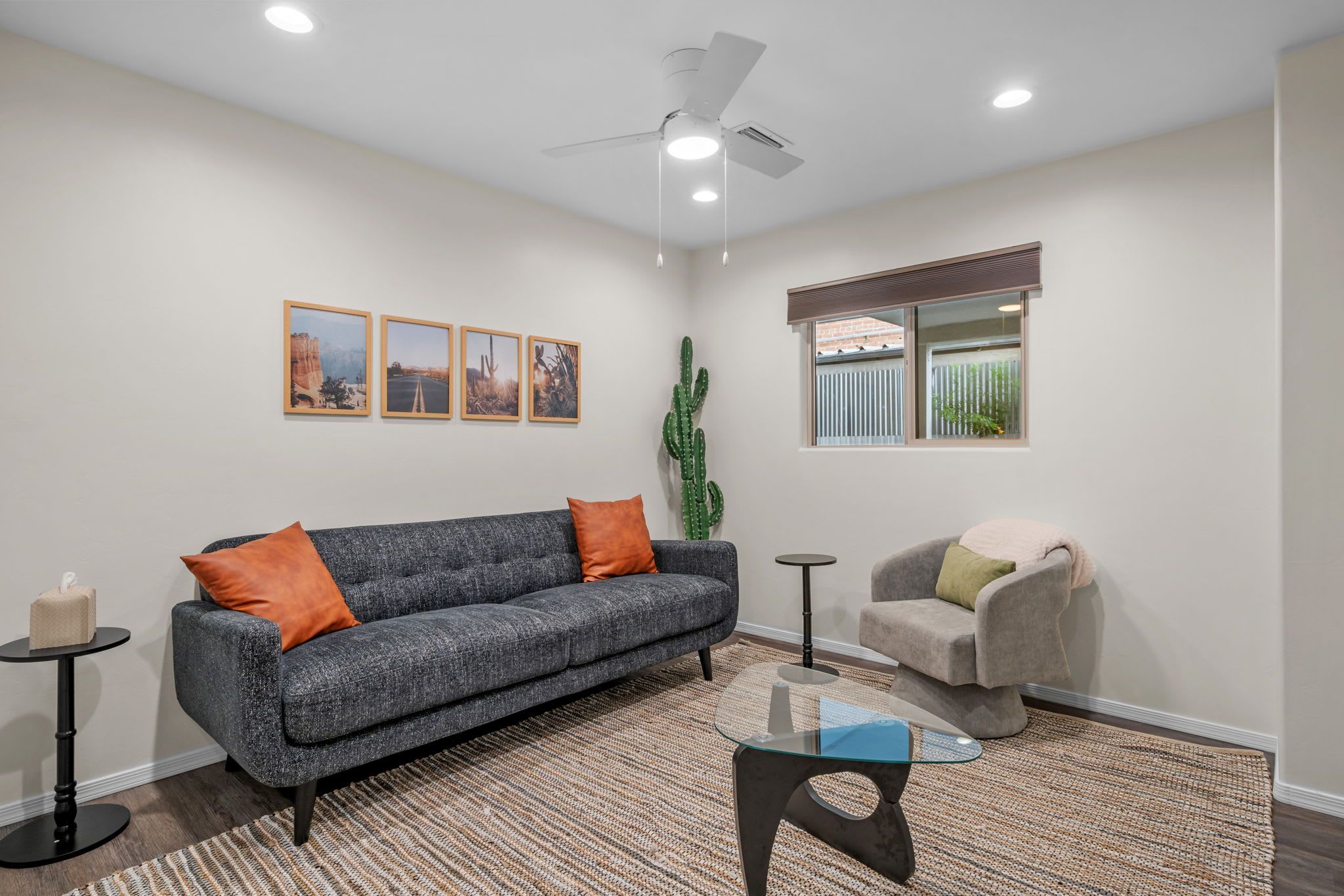 A living room with a grey sofa, orange cushions, a glass coffee table, a grey armchair, a cactus, and framed pictures on the wall.