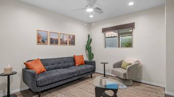 A living room with a grey sofa, orange cushions, a glass coffee table, a grey armchair, a cactus, and framed pictures on the wall.