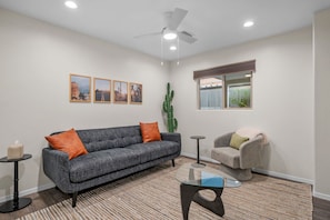 A living room with a grey sofa, orange cushions, a glass coffee table, a grey armchair, a cactus, and framed pictures on the wall. - Presidio Flat at Hoff & Co | Downtown Tucson (Tucson)