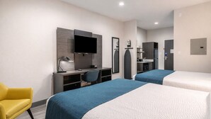 Ein Hotelzimmer mit zwei Betten, einem Schreibtisch mit Stuhl, einem Fernseher, einem Spiegel und einer kleinen Küchenzeile.