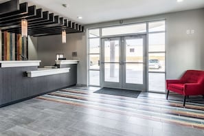 Et receptionsområde med moderne design, der inkluderer en receptionsdisk, en rød stol og en stor glasdør, der fører udendørs.