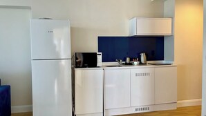 Une petite cuisine équipée avec un réfrigérateur blanc, un four à micro-ondes et un petit plan de travail sur lequel se trouvent une bouilloire et des tasses.