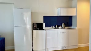 Une petite cuisine équipée avec un réfrigérateur blanc, un four à micro-ondes et un petit plan de travail sur lequel se trouvent une bouilloire et des tasses.